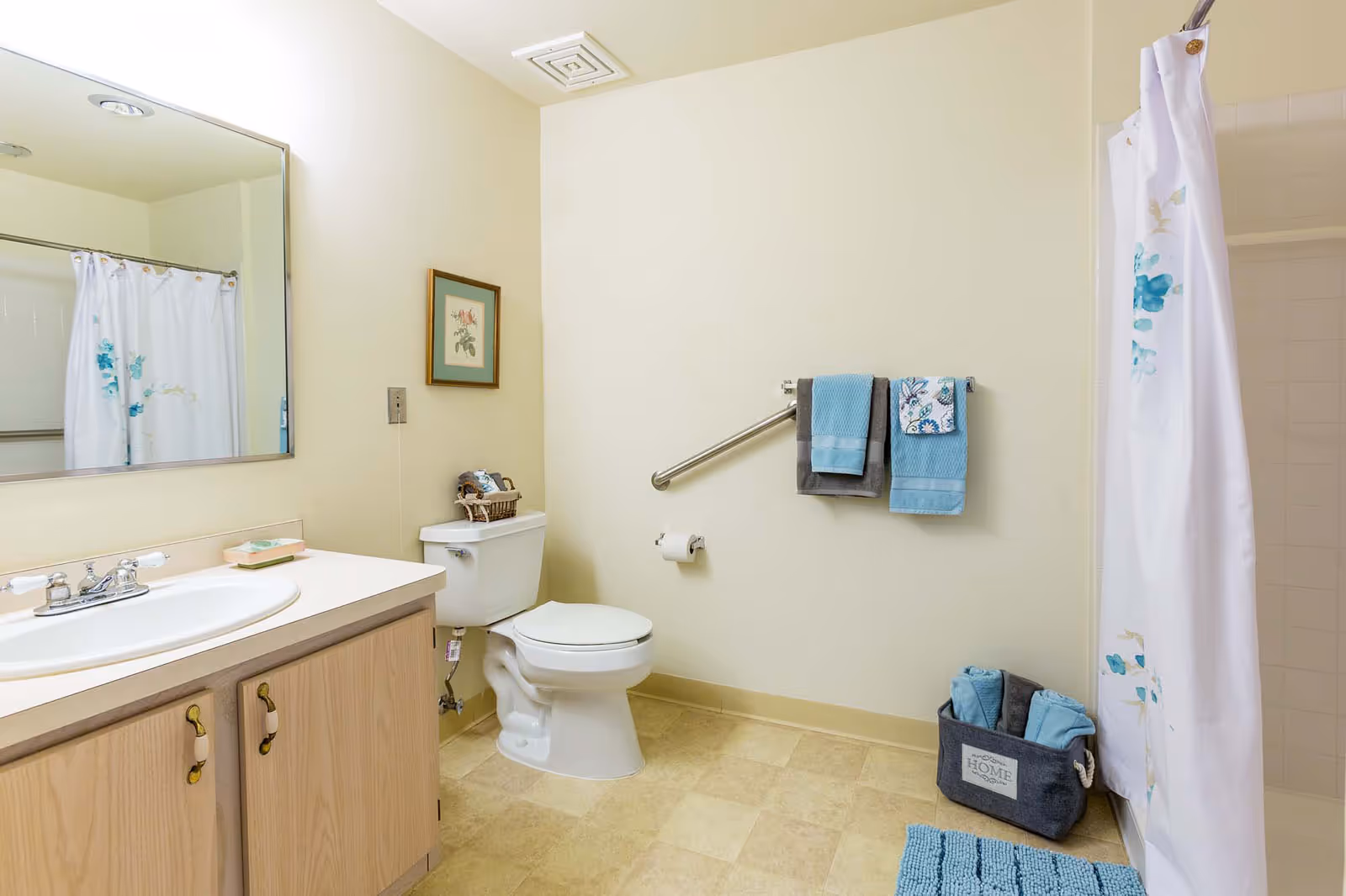 A clean bathroom with a white toilet, a wooden vanity with a white sink, a large mirror above the sink, and a shower with a white curtain featuring blue floral designs. There are blue and gray towels hanging on a metal grab bar on the wall, a basket with towels on the floor, and a framed floral picture on the wall.