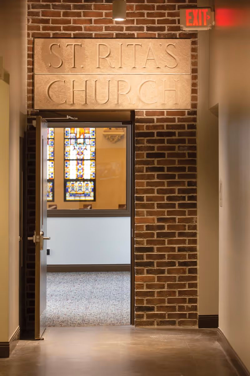 Open doorway in a brick wall with a stone sign above reading 'ST. RITA'S CHURCH'. Through the doorway, stained glass windows and part of the church interior are visible. An illuminated red exit sign is mounted on the wall to the right.