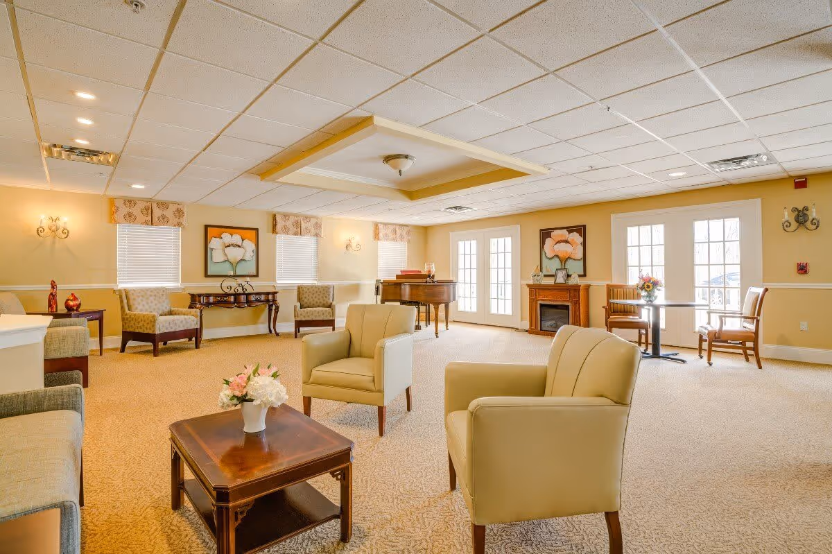 A bright and spacious living room area with beige walls and carpet. The room features comfortable armchairs and sofas arranged around a wooden coffee table with a flower vase. There is a grand piano near the windows, a fireplace with floral artwork above it, and a small dining table with chairs by the French doors letting in natural light.