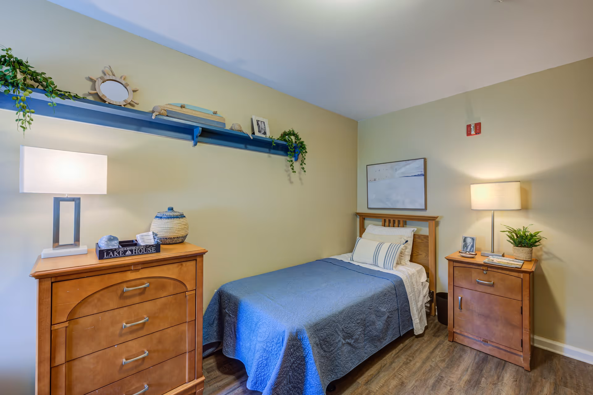 A cozy bedroom with a single bed covered in a blue quilt and white pillows. There are two wooden nightstands on either side of the bed, each with a lamp and decorative items. Above the bed is a framed picture, and a blue shelf on the wall holds plants and small decorative objects. The room has wooden flooring and light-colored walls.