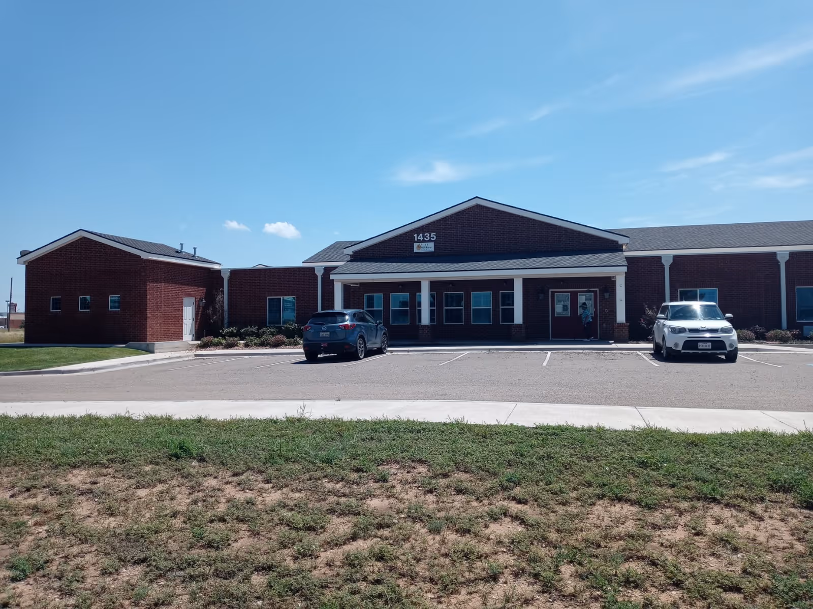Front view of a single-story brick senior living facility with a parking lot and two cars under a clear blue sky.