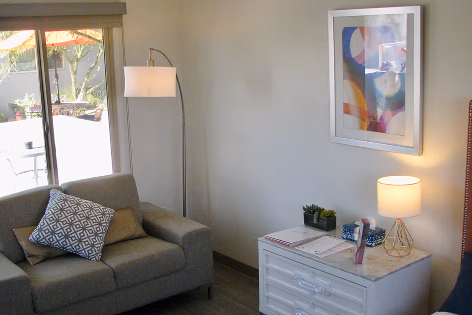 A small living area with a gray sofa and patterned pillow, a floor lamp, a white side table with a lamp and decor, framed wall art, and a sliding glass door to a patio.