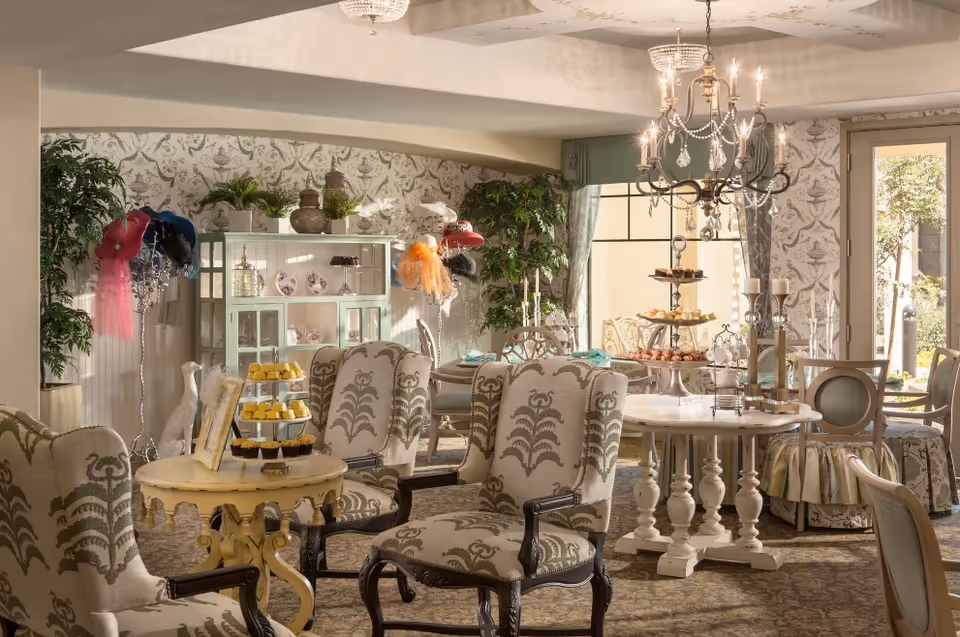 A cozy and elegant interior room with upholstered armchairs and round tables set with tiered trays of pastries and desserts. The room features patterned wallpaper, a chandelier, large windows with curtains, and decorative plants. There is a cabinet with decorative items and hats on display in the background.