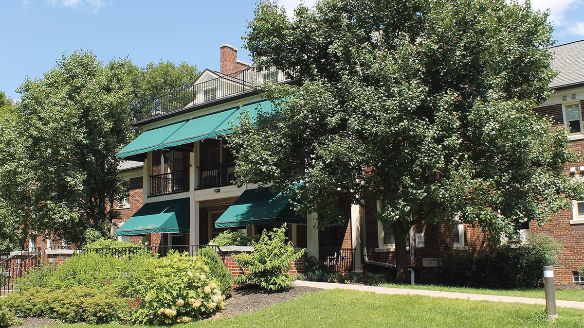 Exterior view of a brick building with green awnings over the windows and balconies, surrounded by trees and well-maintained landscaping under a clear blue sky.