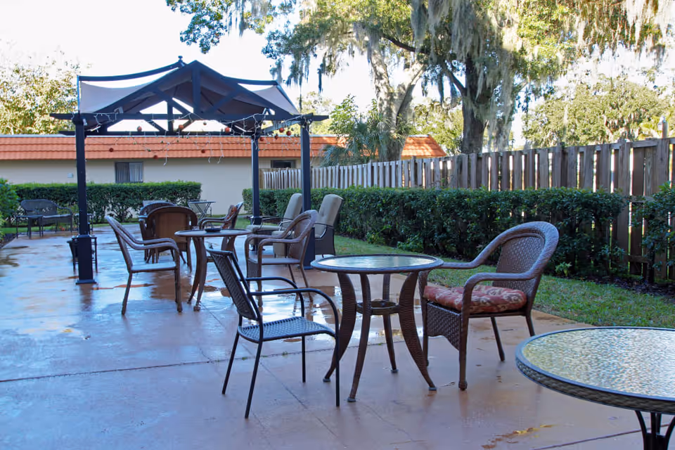 Outdoor patio area with multiple glass-top tables and chairs arranged on a concrete surface. There is a black metal pergola with a canopy and string lights hanging from it. The patio is surrounded by green bushes, a wooden fence, and trees in the background.