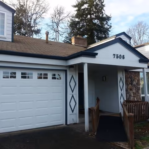 White house-style building with a covered entrance, attached garage, blue trim, and ramped walkway.