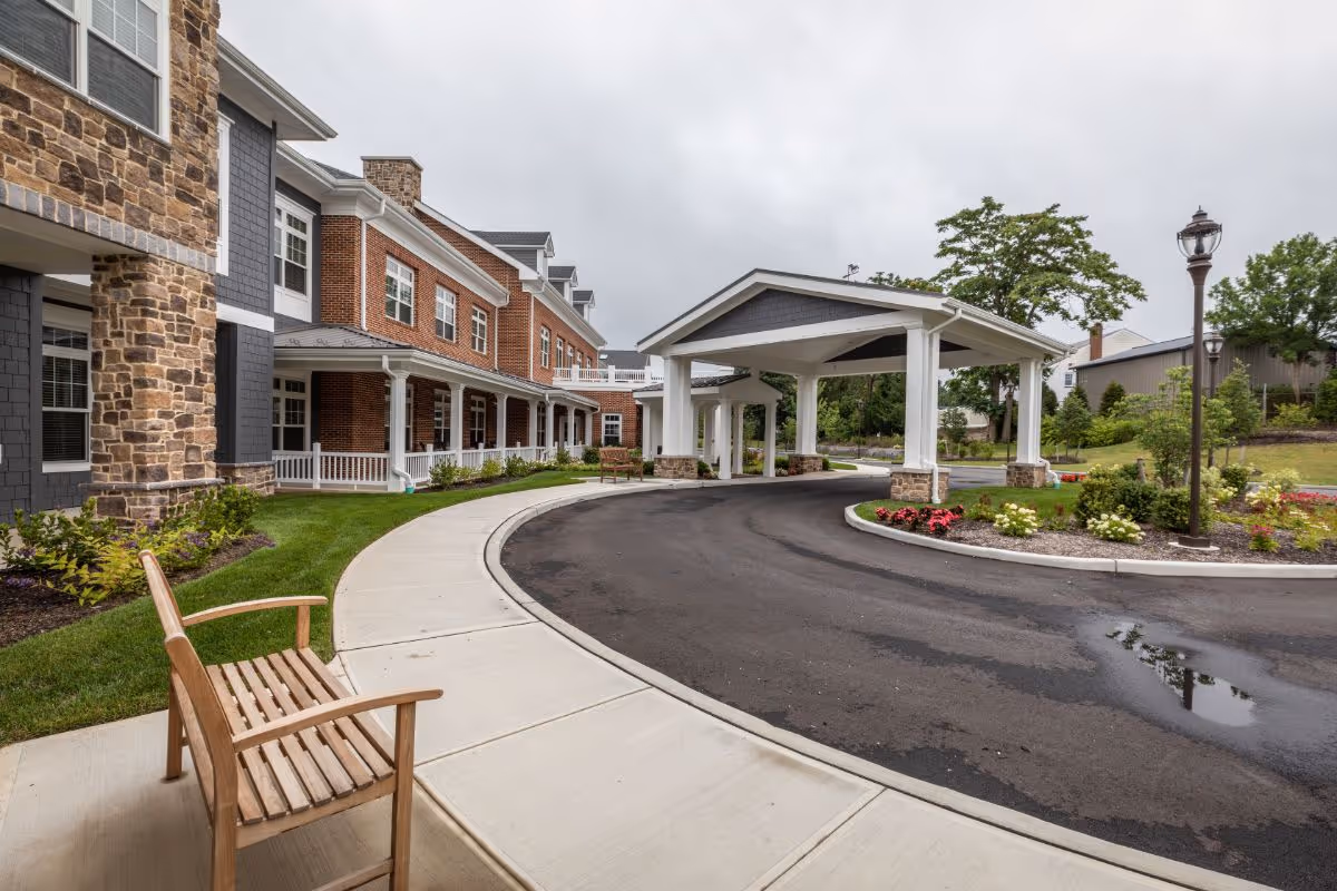 Exterior view of Sunrise of Huntington senior living facility showing a covered driveway entrance, a curved sidewalk, a wooden bench, landscaped flower beds, and a multi-story building with brick and stone facade under a cloudy sky.