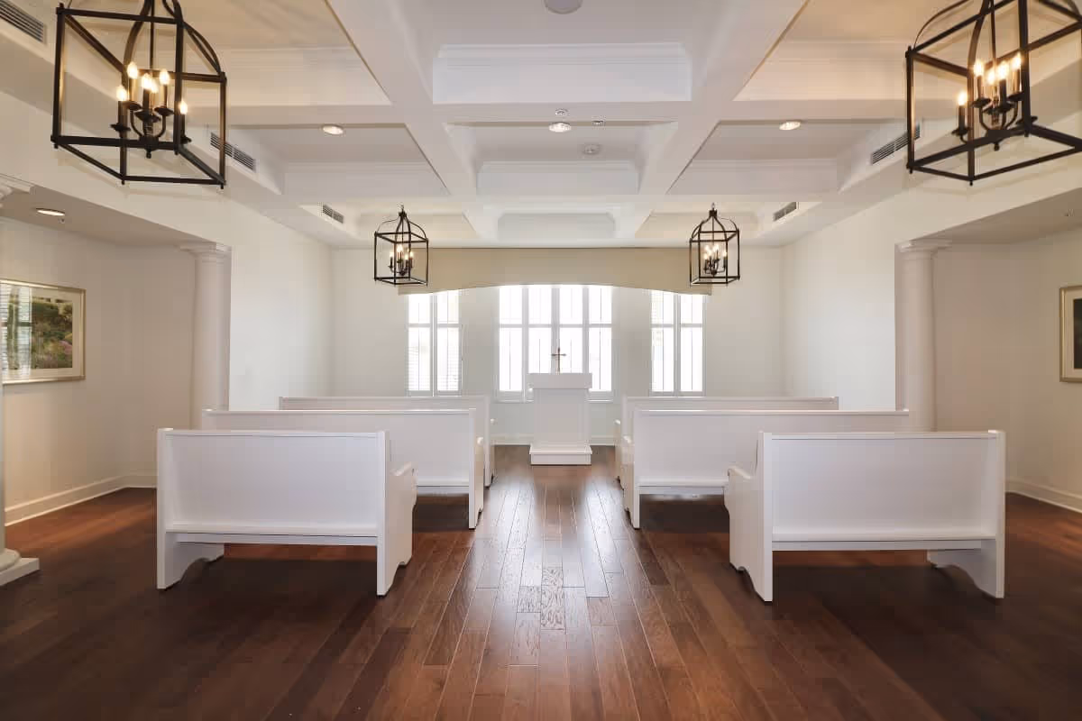 Interior view of a small chapel or meditation room with white wooden pews arranged in two rows facing a white podium with a cross in front of large windows. The room has wooden flooring, white walls, decorative ceiling lights, and framed artwork on the walls.