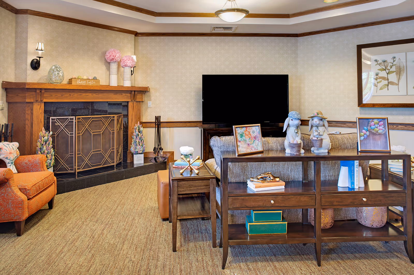A cozy living room with a large flat-screen TV mounted on the wall, a wooden fireplace with decorative items including vases with pink flowers and a 'Happy Easter' sign, an orange armchair, and a wooden console table behind a gray sofa displaying framed pictures and two bunny figurines. The room has beige patterned wallpaper, a carpeted floor, and a ceiling light fixture.