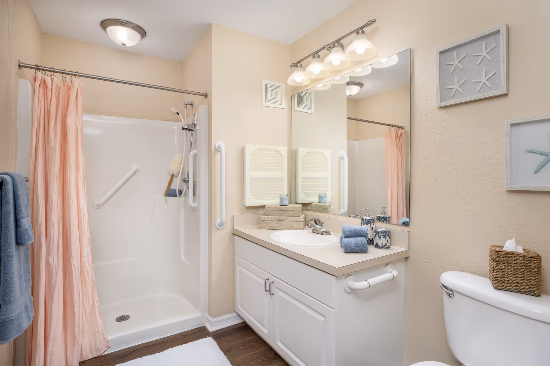 A clean and well-lit bathroom featuring a white shower with a pink curtain, grab bars for safety, a vanity with a sink, a large mirror, and neatly folded blue towels. There is a toilet with a wicker tissue holder on top, and decorative starfish artwork on the beige walls.