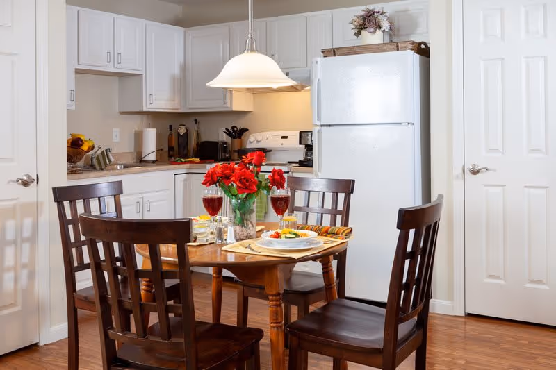 A cozy kitchen and dining area with a wooden table set for two, featuring plates of food, glasses of red wine, and a vase of red flowers. The kitchen has white cabinets, a white refrigerator, a stove, and various kitchen appliances. The floor is wooden, and there are two closed white doors visible.