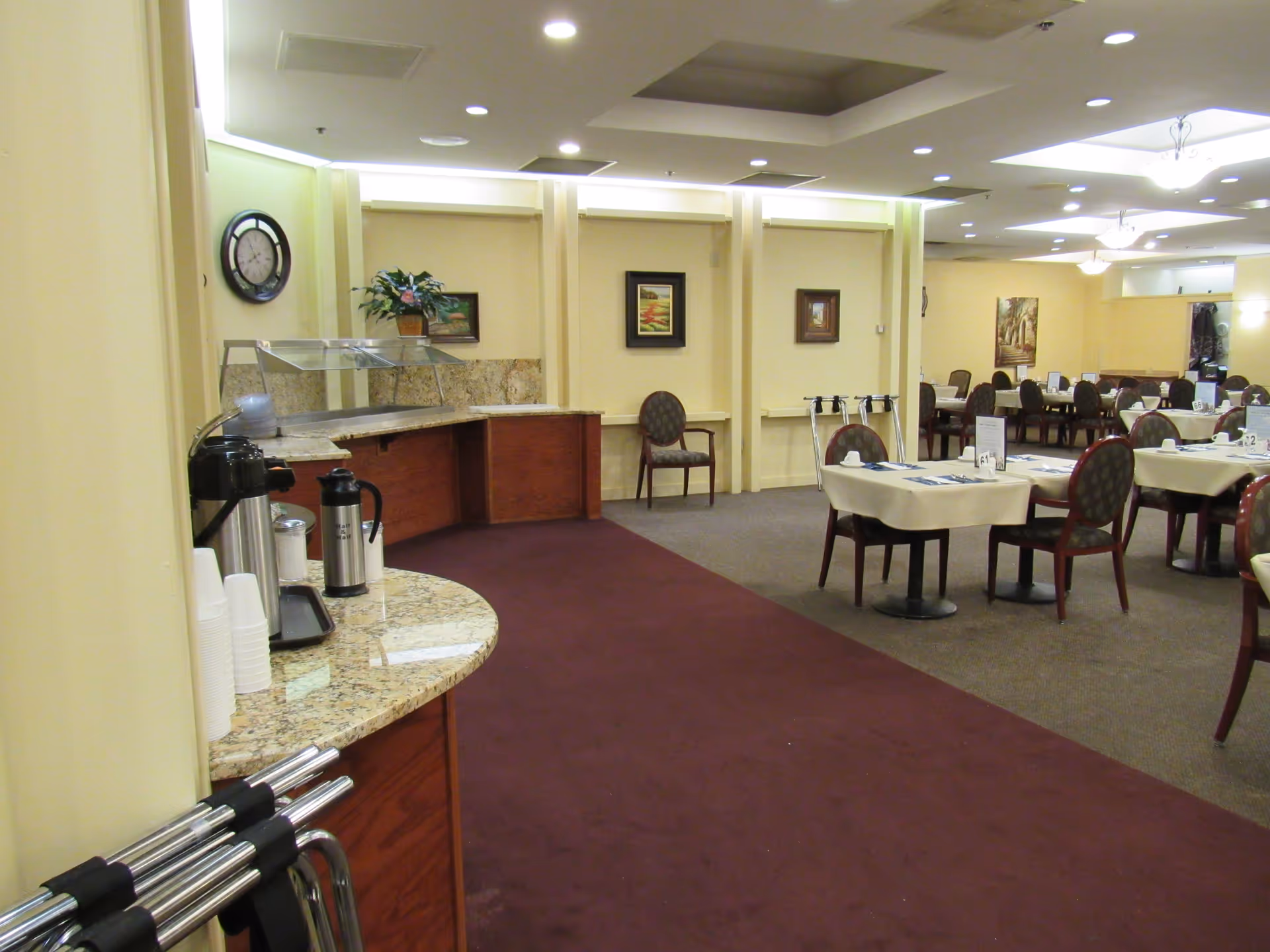 A dining room with set tables and chairs, a buffet counter and coffee station in a senior living facility.