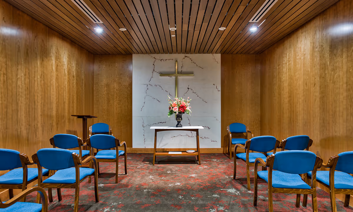 A small chapel room with wooden paneled walls and ceiling, featuring a gold cross mounted on a marble wall at the front. Below the cross is a table with a vase of colorful flowers. The room has blue cushioned wooden chairs arranged in rows facing the front.