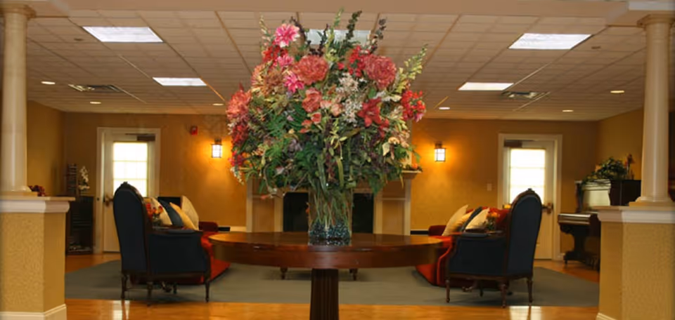A cozy senior living facility common area with a large round wooden table in the center holding a tall vase filled with a colorful bouquet of flowers. The room has warm yellow walls, two dark blue armchairs with cushions, a red sofa, and two doors with windows. There are two columns on either side of the room and ceiling lights providing illumination.