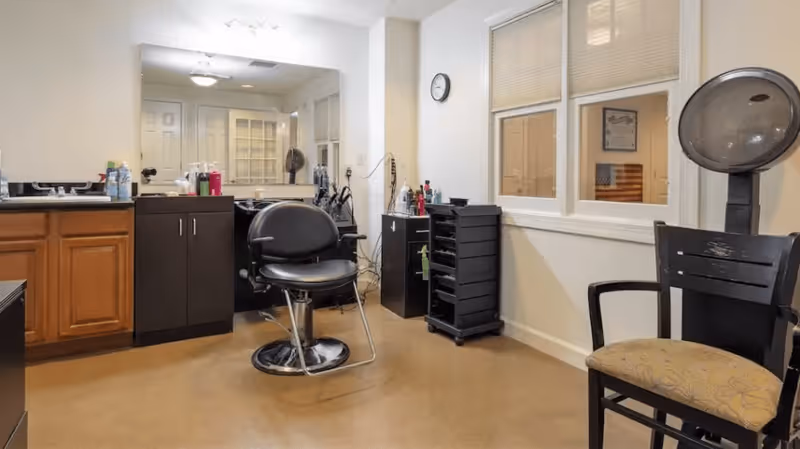 Interior view of a salon area in a senior living facility with a black salon chair in front of a large mirror, wooden cabinets, a hair dryer chair, and various hair styling tools and products on counters and shelves.