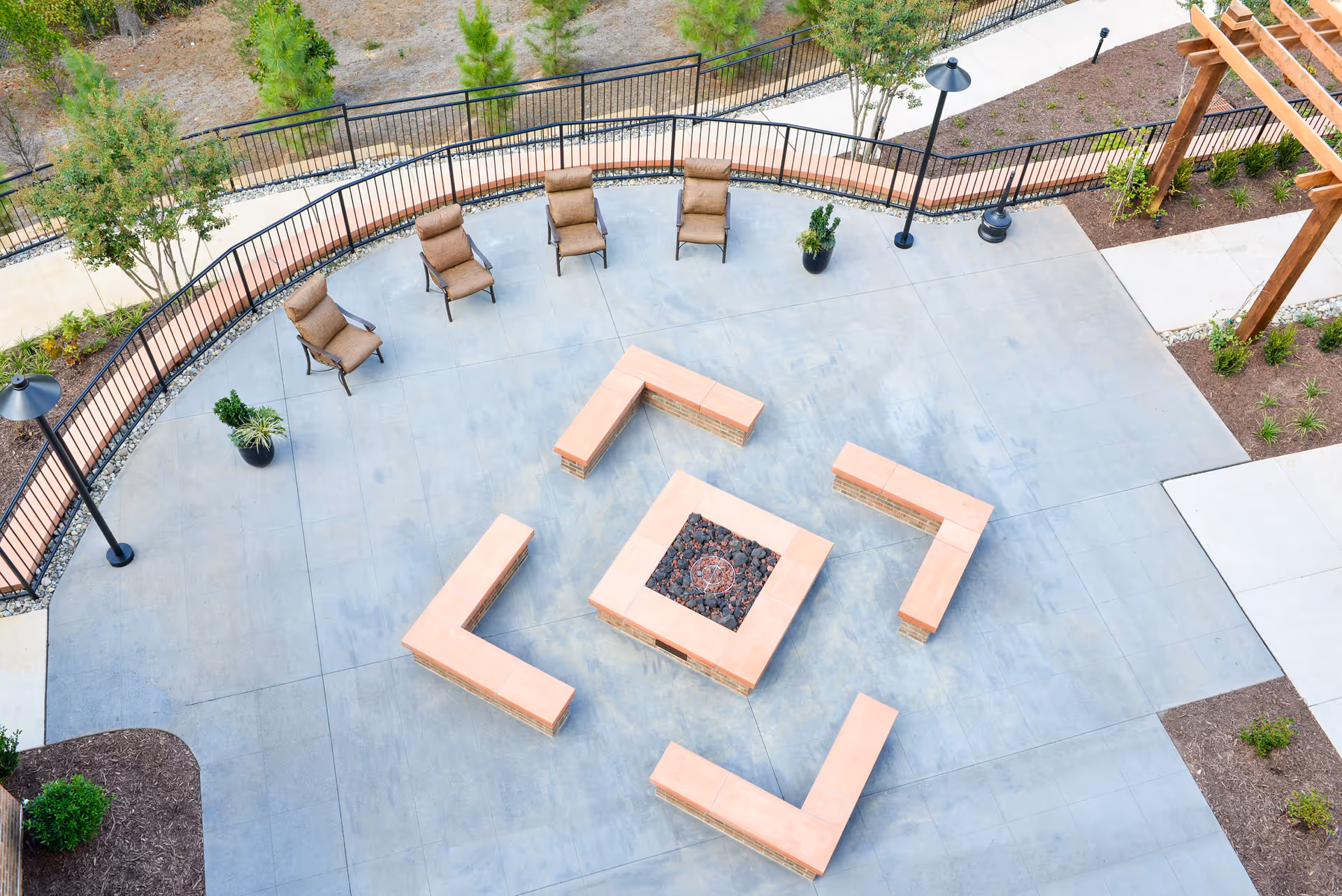 Aerial view of an outdoor courtyard with a central square fire pit surrounded by L-shaped benches, lounge chairs, potted plants, lampposts and a pergola.