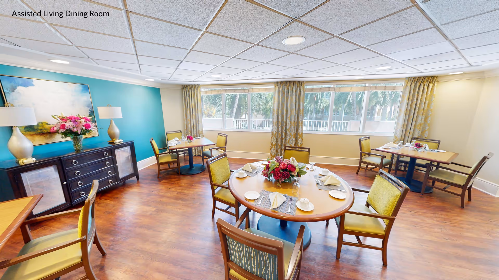 Bright assisted living dining room with wooden floors, round and square tables set with white napkins, cups, and plates. Chairs with green cushions surround the tables. A sideboard with two lamps and a vase of flowers is against a teal accent wall. Large windows with patterned curtains let in natural light and show greenery outside.