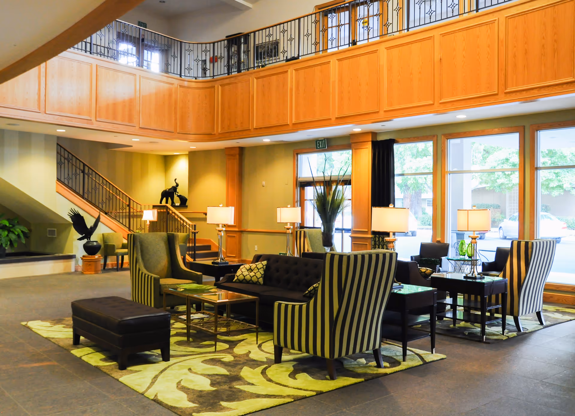 A spacious and well-lit living room area with a high ceiling and wooden balcony railing above. The room features a black tufted sofa, striped armchairs, a black ottoman, glass-top coffee tables, and several table lamps. Large windows allow natural light to fill the space, and decorative sculptures and plants are placed near a staircase in the background.