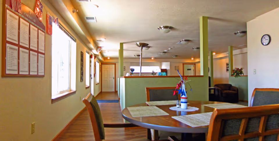 Interior view of a senior living facility common area with a round wooden table set with placemats and a patriotic-themed centerpiece. The room has wooden flooring, light-colored walls, several windows letting in natural light, and green half-walls dividing the space. There are chairs around the table and a comfortable armchair in the background. A clock is mounted on the wall.
