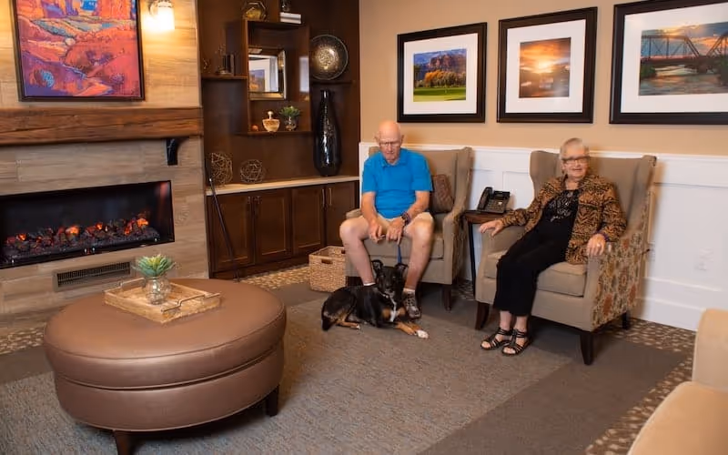 A cozy living room with a fireplace, a round brown ottoman, and two elderly people sitting in armchairs. A black and brown dog is lying on the carpet between them. The wall behind them has three framed landscape photographs, and there is a wooden cabinet with decorative items to the left.