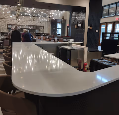 Interior view of a modern dining area with a large white curved countertop bar in the foreground, brown chairs around the bar, and tables and chairs in the background. Several people are standing near the back of the room. The ceiling has decorative hanging lights and the walls feature a mix of stone and dark paneling.