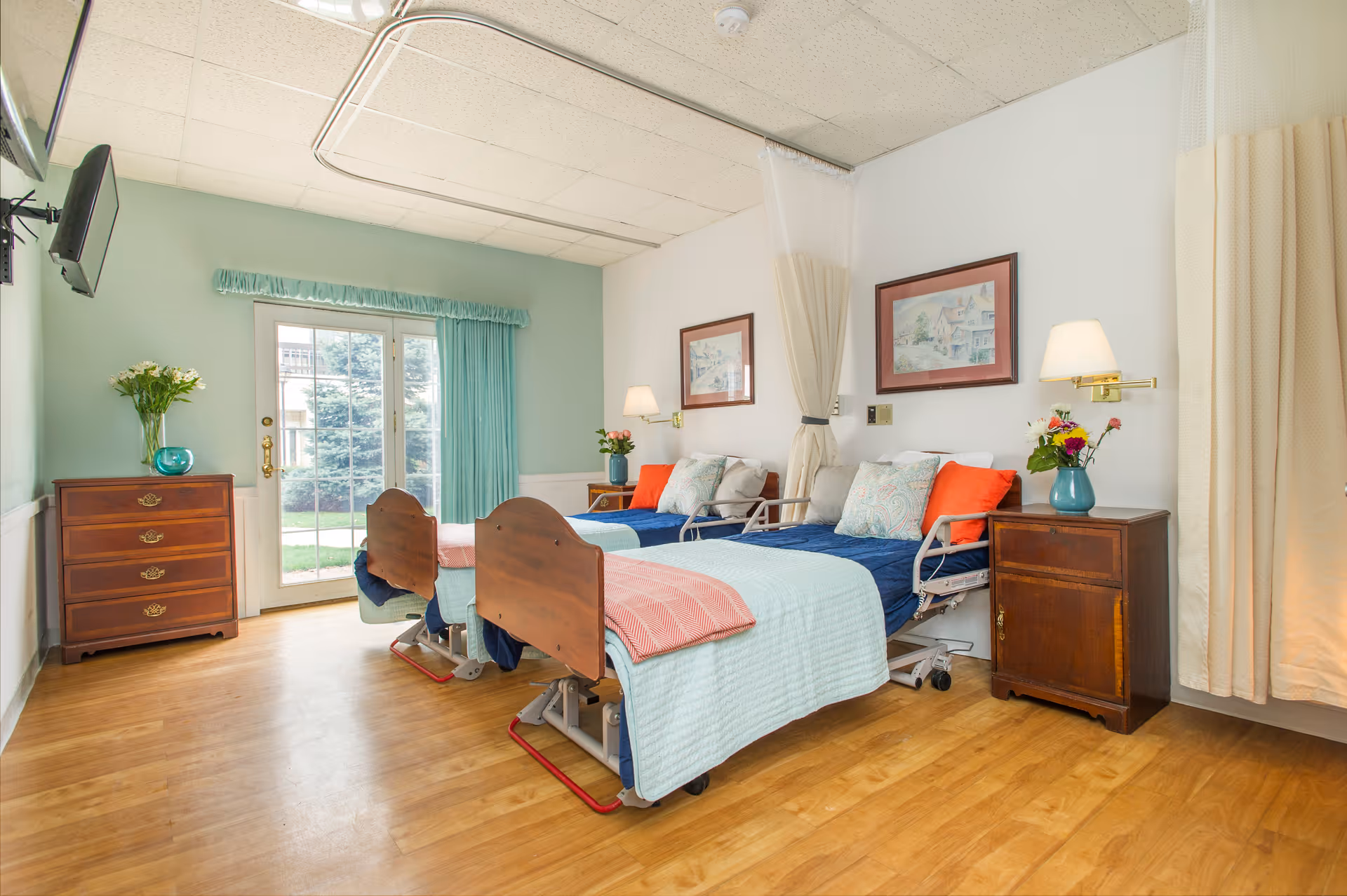 A bright and clean bedroom in a senior living facility with two hospital-style beds, each made with blue and light blue bedding and orange accent pillows. The room features wooden furniture including a chest of drawers and a nightstand with vases of flowers. There are framed pictures on the walls, a wall-mounted TV, and large glass doors with teal curtains leading outside. The floor is wooden, and there are privacy curtains between the beds.