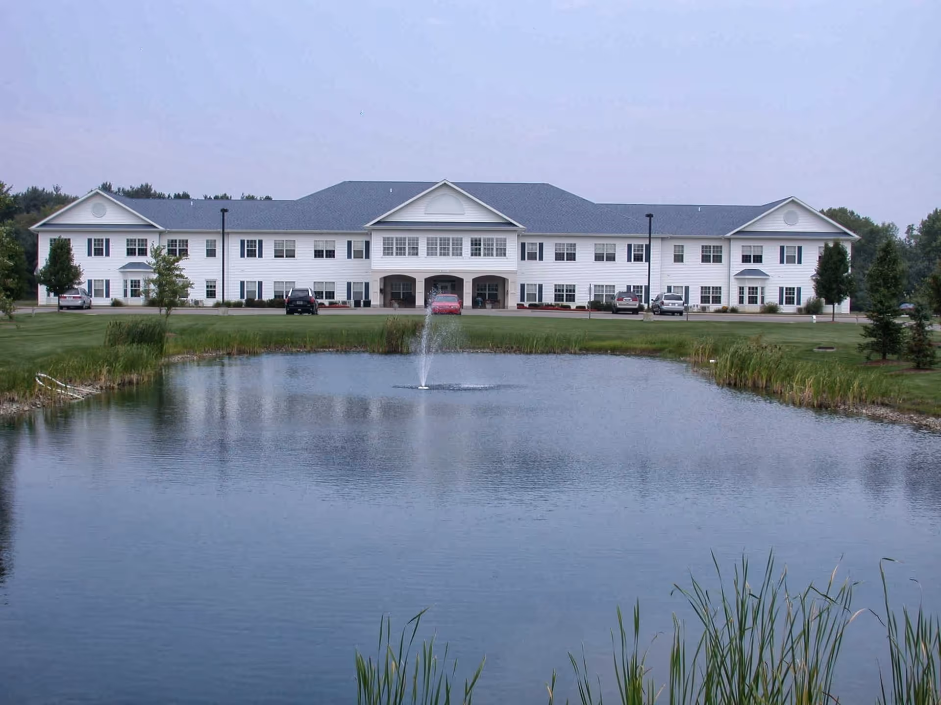 White two-story residential building with a central entrance and parked cars, reflected across a pond with a fountain.