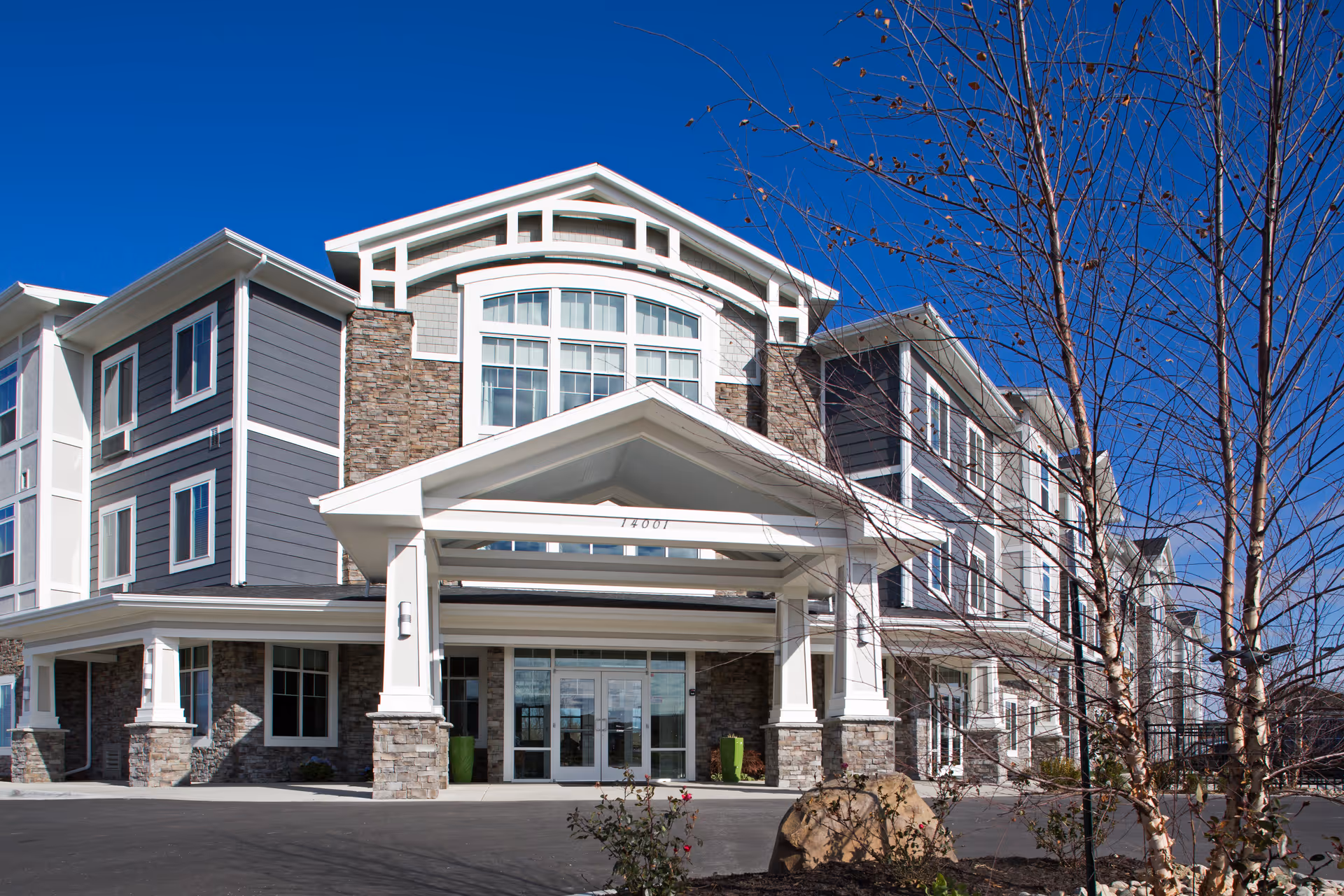 Exterior front view of a modern three-story senior living facility with stone and gray siding, large windows, and a covered entrance under a clear blue sky.