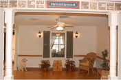 Interior view of a room with white walls and wooden floor, featuring a ceiling fan with light, two wall-mounted lantern-style lights, a window with green shutters and white curtains, a wicker chair with a cushion, two potted plants, a small wooden chest, and a decorative dog statue. Above the room entrance is a sign that reads 'Brentwood'.