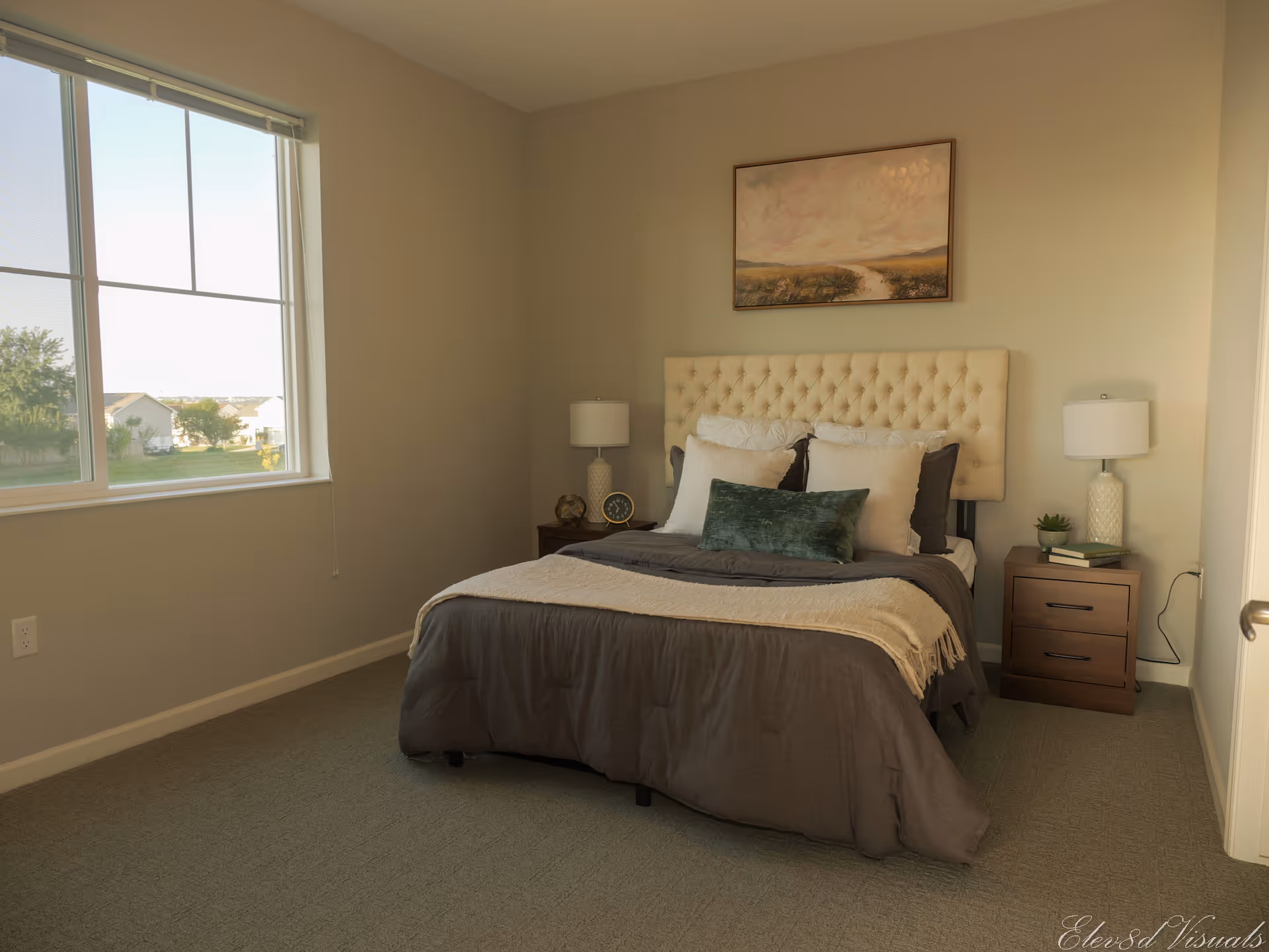 A neatly made bed with a tufted beige headboard in a bedroom. The bed has multiple pillows and a dark gray comforter with a light beige throw blanket. On either side of the bed are wooden nightstands with white lamps, a clock, books, and a small plant. A large window on the left side lets in natural light, and a landscape painting hangs above the bed.