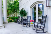 Front porch with a row of black rocking chairs, potted plants, white columns, and arched windows overlooking a garden.
