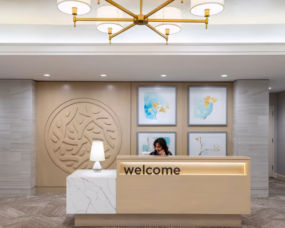Bright reception area with a receptionist behind a light wood desk reading 'welcome' and decorative wall art behind it.