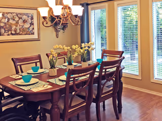 A dining room with a wooden table set for six people, featuring placemats, turquoise bowls, and three small potted plants as a centerpiece. The room has large windows with white blinds and a chandelier hanging above the table. A framed floral artwork is on the wall.