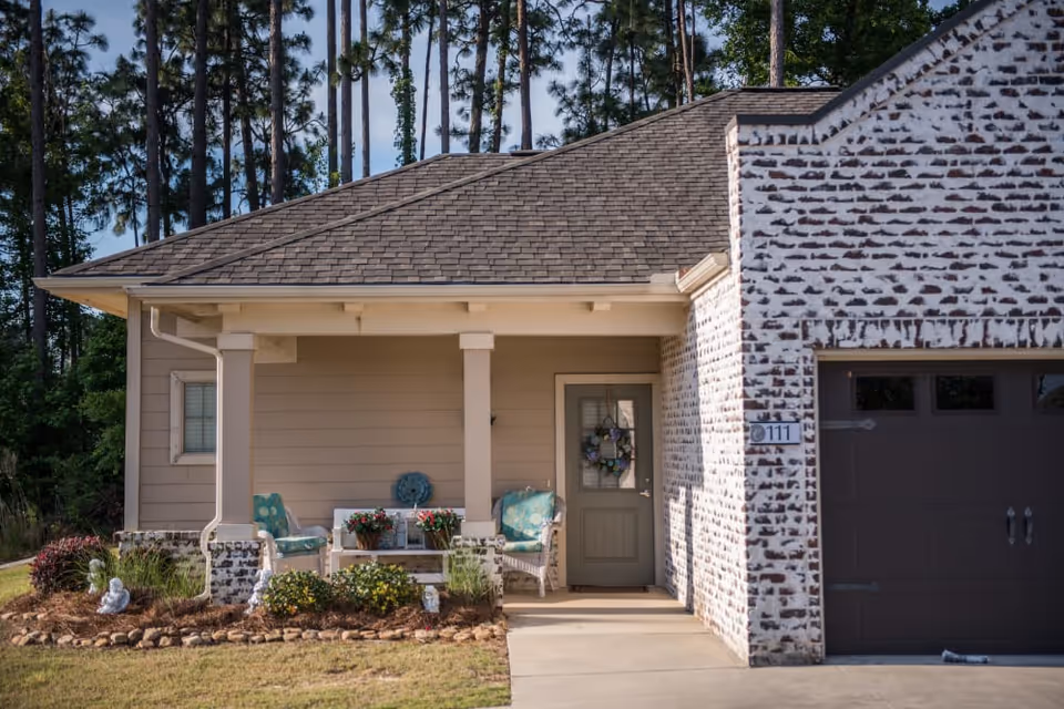Front porch of a residential unit with a small seating area including two chairs and a table with flower pots. The house has beige siding and a brick section near the garage door. There is a wreath on the front door and tall pine trees in the background.