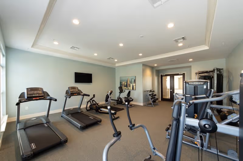 A senior living facility fitness room with exercise equipment including treadmills, stationary bikes, weight machines, and free weights. The room has light blue walls, recessed ceiling lights, a wall-mounted TV, and large windows letting in natural light.