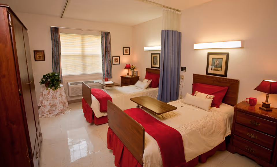 Two neatly made twin beds with red accents in a senior living bedroom, separated by a privacy curtain, with bedside tables, wall lights and a window air conditioner.