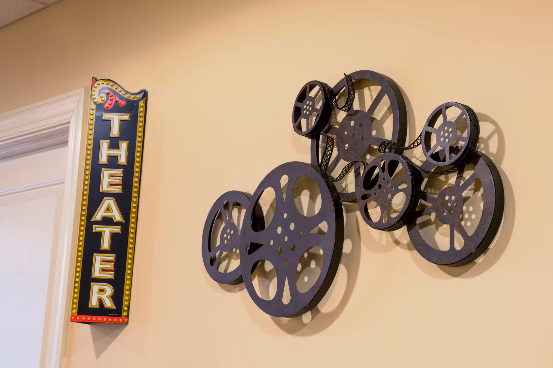 Wall decoration featuring a vertical sign with the word 'THEATER' in yellow letters on a black background with a decorative border, next to a metal wall art piece depicting several film reels with film strips.
