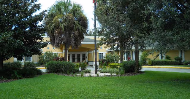 Front exterior view of a single-story yellow building with a covered entrance, surrounded by green grass, bushes, and trees including a palm tree and other leafy trees.