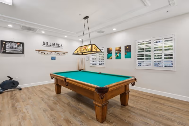 A bright room with a wooden billiards table featuring a green felt surface. Above the table hangs a decorative stained glass light fixture. The walls are white with several billiards-themed pictures and a sign that reads 'BILLIARDS'. There are two windows with white shutters letting in natural light, and a stationary exercise bike is positioned in the corner. The floor is covered with light wood laminate.