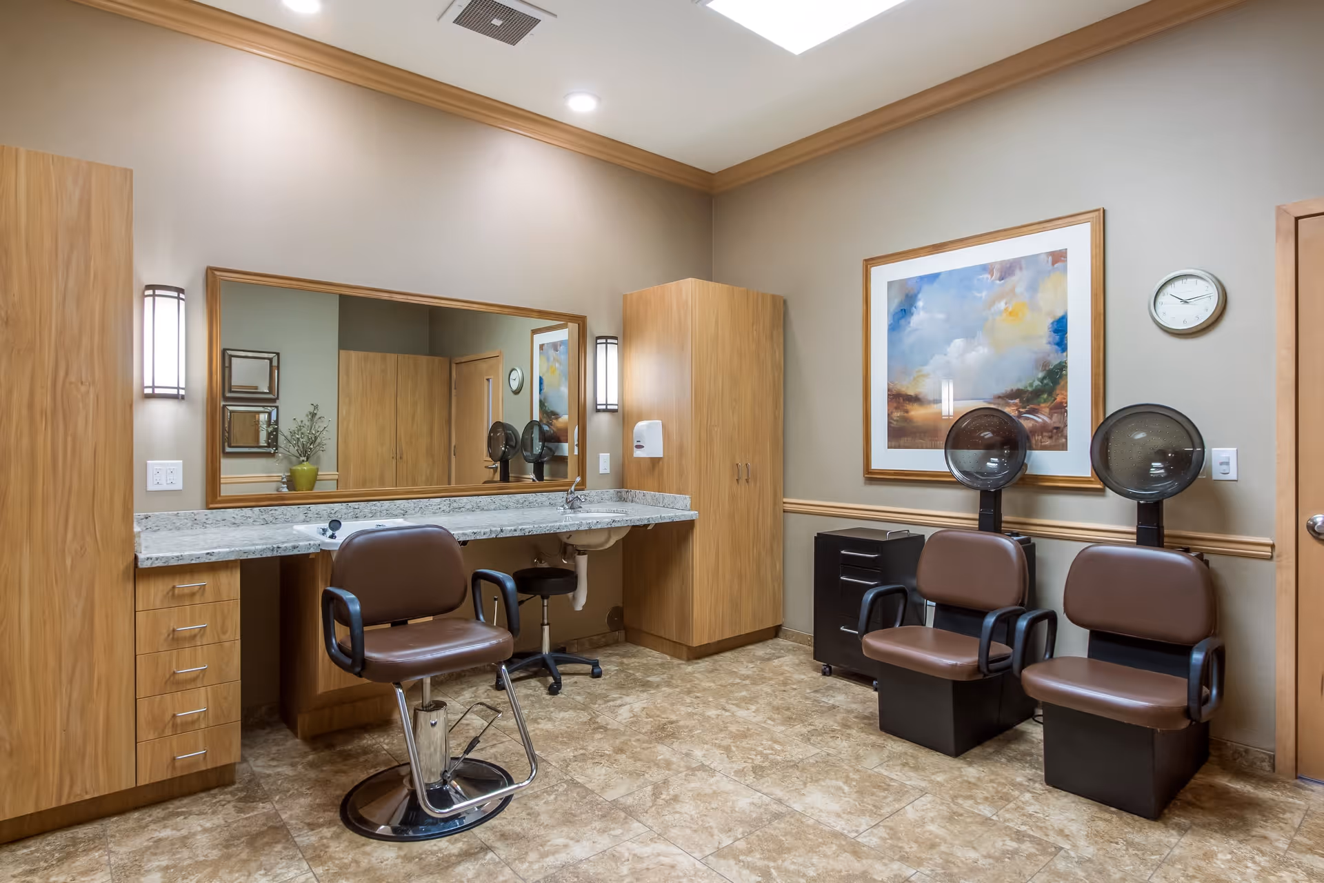 Interior of a salon area in a senior living facility with two brown salon chairs under hair dryers, a large mirror above a granite countertop with a salon chair in front, wooden cabinets, a framed painting on the wall, and a clock.