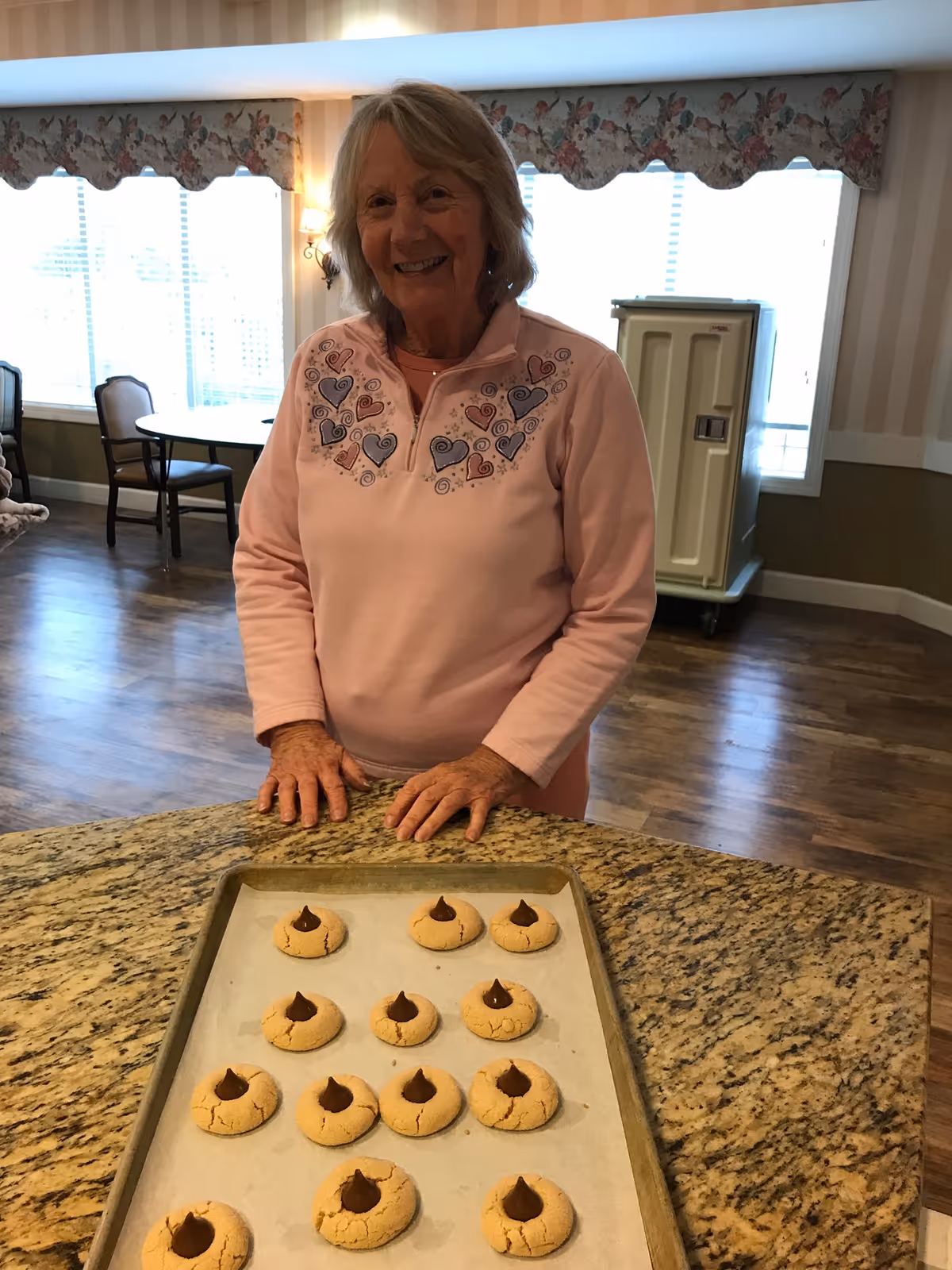 An elderly woman wearing a pink sweater with heart designs stands behind a kitchen counter with a baking sheet of freshly baked cookies topped with chocolate kisses. The room has wooden floors, large windows with floral valances, and several chairs and tables in the background.