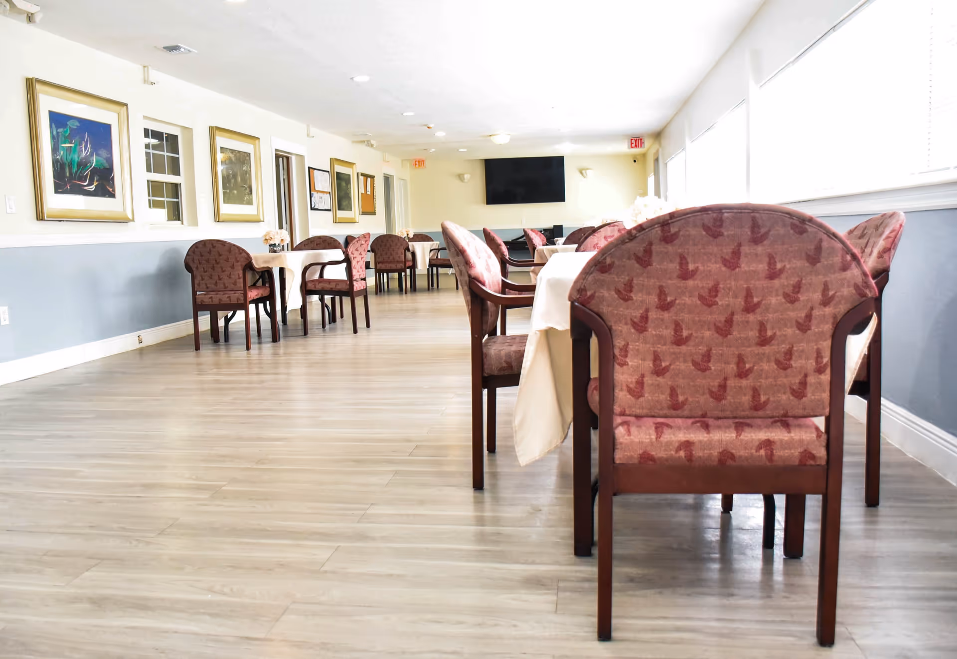 A bright dining room with multiple tables covered with white tablecloths and surrounded by maroon upholstered chairs. The room has light wood flooring, light-colored walls with framed artwork, and large windows on one side letting in natural light. A flat-screen TV is mounted on the far wall.