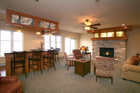 A cozy living room area featuring a stone fireplace with a lit fire, surrounded by comfortable armchairs and a sofa. There is a wooden coffee table in the center, a side table with a lamp, and a bar counter with high chairs near large windows letting in natural light. The ceiling has a fan with lights, and the room has a warm, inviting atmosphere.