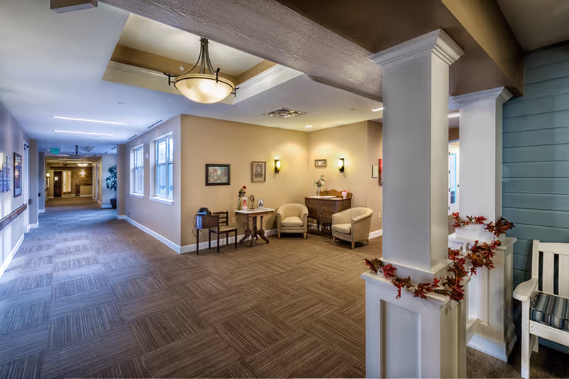 Carpeted interior hallway and sitting area with armchairs, side tables, and overhead lighting in a senior living facility.