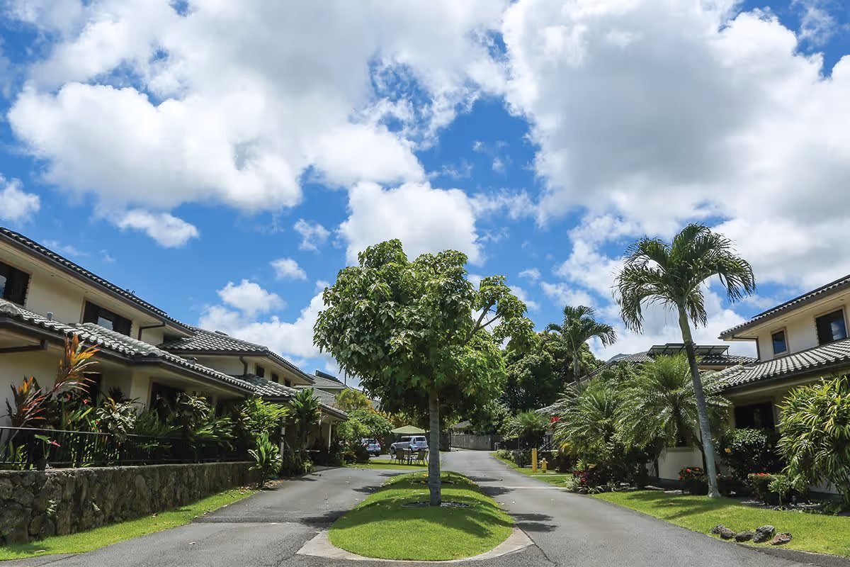 A sunny outdoor view of Kina 'Ole Estate showing a paved driveway split by a small grassy island with a tree in the center. The driveway is flanked by two-story residential buildings with tiled roofs and lush tropical landscaping including palm trees and various shrubs. The sky is bright blue with scattered white clouds.