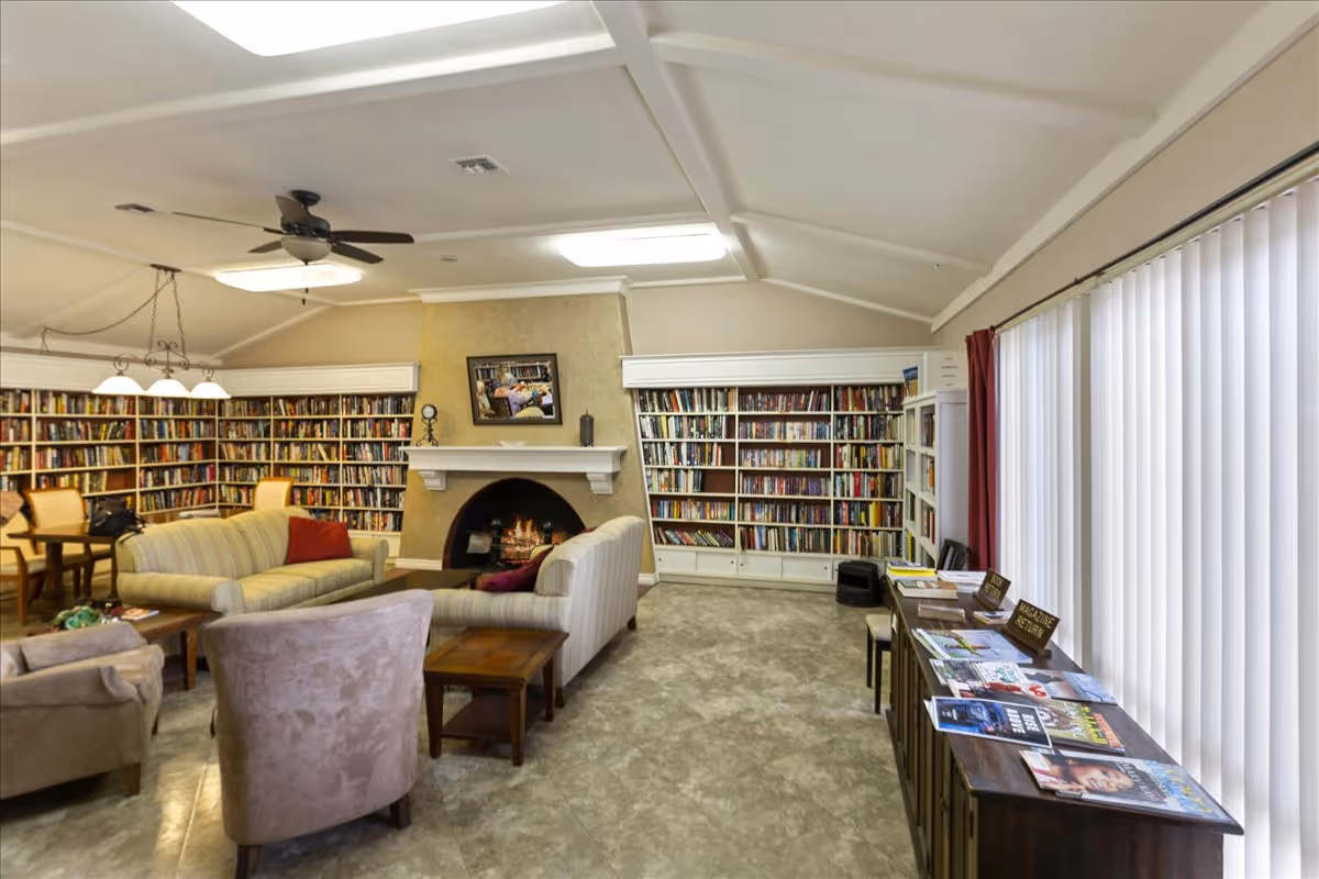 Cozy communal library lounge with bookshelves lining the walls, a fireplace, sofas and chairs, and reading tables near vertical blinds.