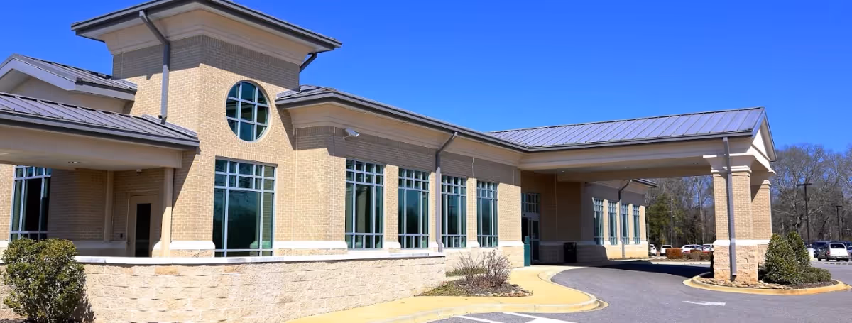 Exterior view of Bibb Med Center Nursing Home building with beige brick walls, multiple large windows, a covered entrance with columns, and a clear blue sky.
