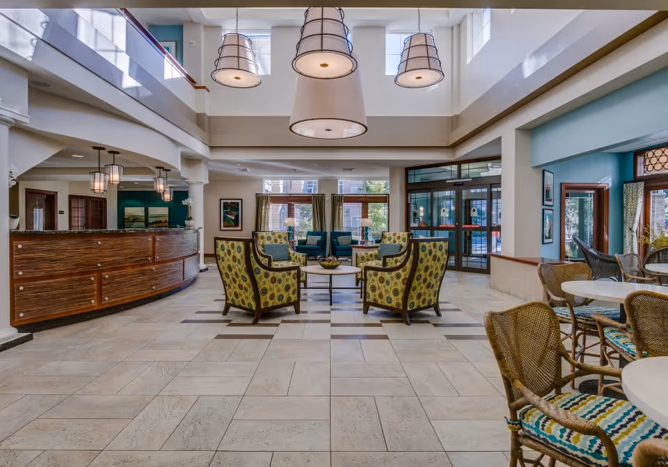 Spacious and well-lit senior living facility lobby with a high ceiling and large hanging light fixtures. The seating area includes patterned armchairs arranged around a coffee table, with additional wicker chairs and tables on the right. A curved wooden reception desk is on the left, and large windows and glass doors provide natural light and an outdoor view.