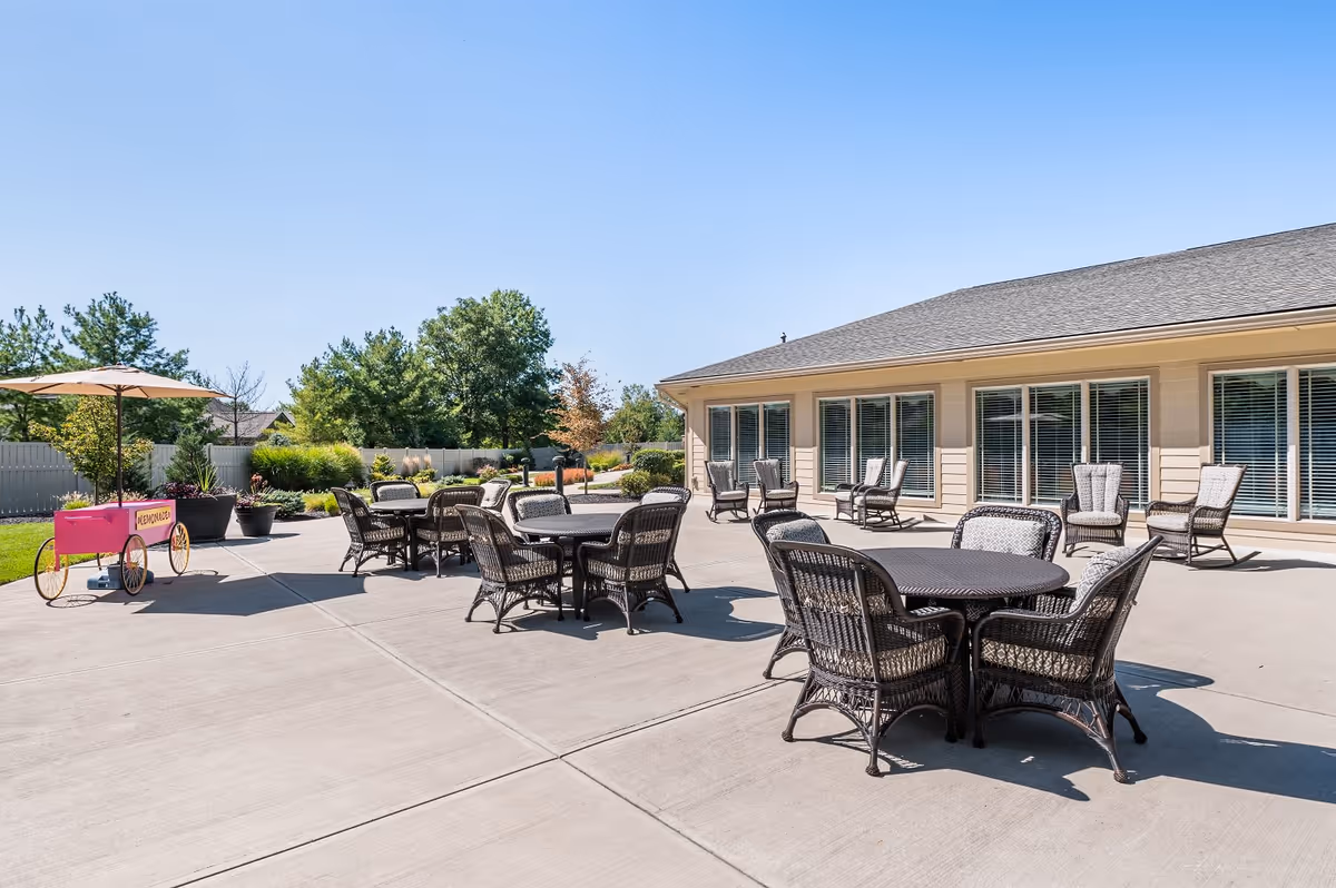 Outdoor patio area at Sage Park Assisted Living & Memory Care with several round tables and wicker chairs arranged on a concrete surface. There is a pink lemonade cart with an umbrella on the left side and a building with large windows and additional chairs in the background. Trees and greenery surround the fenced area under a clear blue sky.