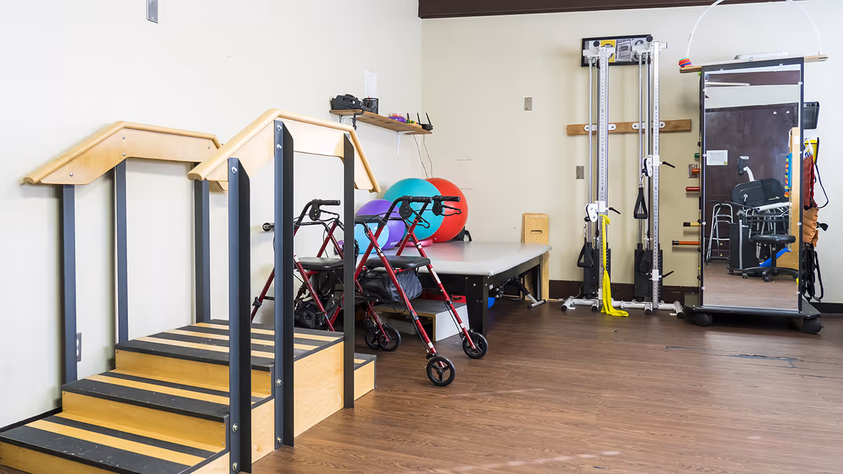 A physical therapy or rehabilitation room with wooden stairs with handrails, two red walkers, a therapy table with colorful exercise balls behind it, a tall exercise machine, and a large mirror on wheels reflecting part of the room.